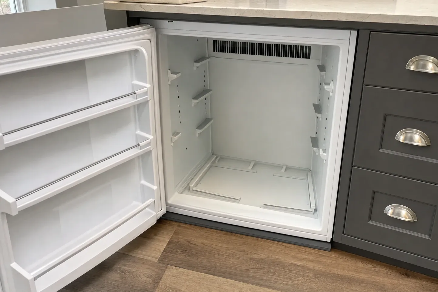 Undercounter refrigerator open in a run of dark shaker cabinets, empty liner with a vent grille along the top