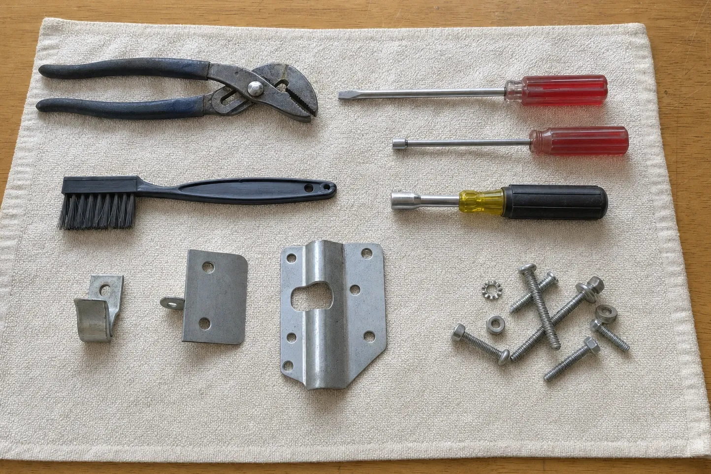 Slip-joint pliers, a wire brush, three nut drivers, hinge brackets and assorted bolts laid out on a cloth